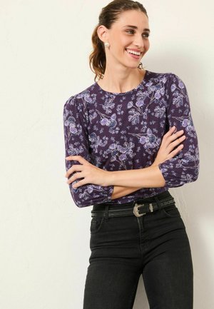 Lila Langarmshirt mit floralem Druck in Hellblau und Lila, gerippter Textur und Rundhalsausschnitt, kombiniert mit schwarzen Jeans und einem Gürtel.