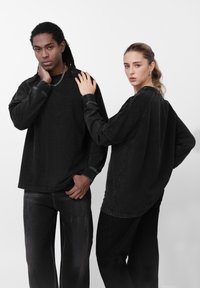 Tops negros de gran tamaño con mangas largas y vaqueros de pierna ancha, con textura desgastada y costuras en contraste, llevados por dos modelos de espaldas.