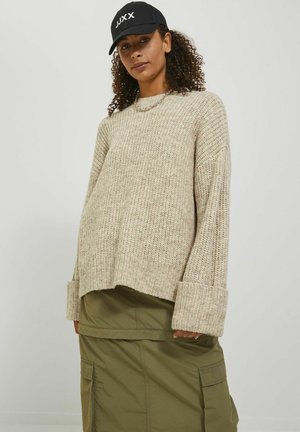 Beige gebreide trui met brede mouwen en geribbelde manchetten, gecombineerd met een olijfgroene rok met zakken. Een zwarte pet maakt de outfit compleet.