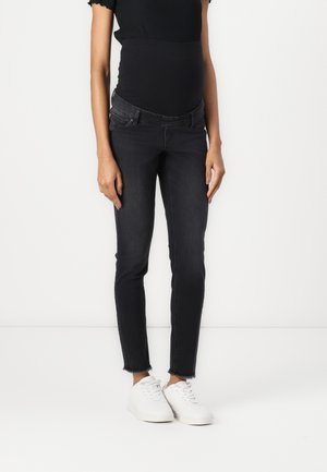 Woman wearing black fitted maternity jeans, black top, and white sneakers, standing against a plain white background.