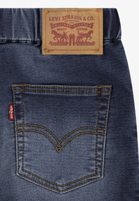 Vaqueros de mezclilla oscura con un diseño de bolsillo trasero cosido en amarillo en forma de "V" distintiva y una etiqueta de cuero marrón con la marca en la cinturilla.