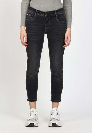 Slim-fit black jeans on a person wearing a grey ribbed long-sleeve top and white sneakers, standing against a white background.