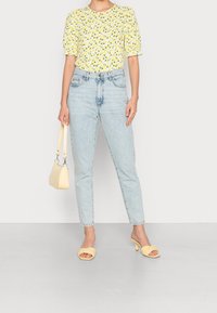 Kobieta w żółtej bluzce z bufkami w wzór cytrynowy, jasnoniebieskich jeansach z wysokim stanem, żółtych sandałach na obcasie i trzymająca małą żółtą torebkę.