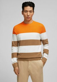 Strickpullover mit Rundhalsausschnitt, der orange, weiß und braun gestreift ist, mit strukturiertem Gewebe und gerippten Bündchen. Getragen mit hellbeigen Hosen.