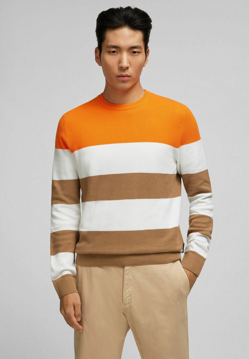 Strickpullover mit Rundhalsausschnitt, der orange, weiß und braun gestreift ist, mit strukturiertem Gewebe und gerippten Bündchen. Getragen mit hellbeigen Hosen.
