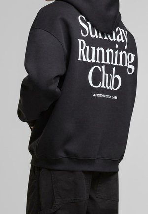 Person trägt einen schwarzen Hoodie mit weißem Text „Sunday Running Club“ und kleinerem Text „Another Cotton Lab“ auf dem Rücken, vor grauem Hintergrund.