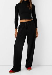 Haut court noir à manches longues avec un col montant et une fermeture éclair, associé à un pantalon noir à jambes larges. Chaussures rouges avec une semelle texturée.