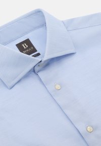 Camicia tessuta di colore azzurro chiaro con colletto strutturato, dotata di due bottoni bianchi e un'etichetta nera con testo bianco.