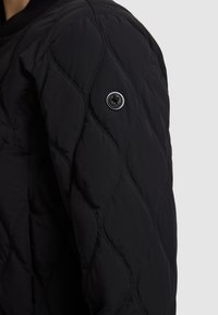 Manche d'une veste matelassée noire avec des piqûres en forme de losange et un badge métallique rond avec un logo en forme de feuille sur le haut du bras.