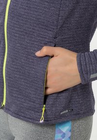 Lila zip-up jacka med en texturerad yta, med en gul dragkedja och sidoficka, tillsammans med grå byxor som visar ett geometriskt mönster.