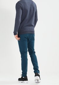 Felpa blu marina con collo a giro, abbinata a pantaloni slim fit verde acqua e sneakers nere con suole bianche. Texture liscia e vestibilità casual.