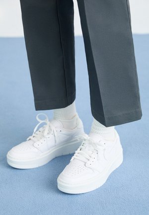 Białe skórzane sneakersy z perforowanym noskiem, płaskimi sznurówkami i grubą białą podeszwą, noszone z szarymi spodniami i prążkowanymi białymi skarpetami.