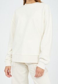 Krémová mikina vyrobená z měkkého materiálu, s dlouhými rukávy, kulatým výstřihem a volným střihem. Bezešvé manžety a lem.