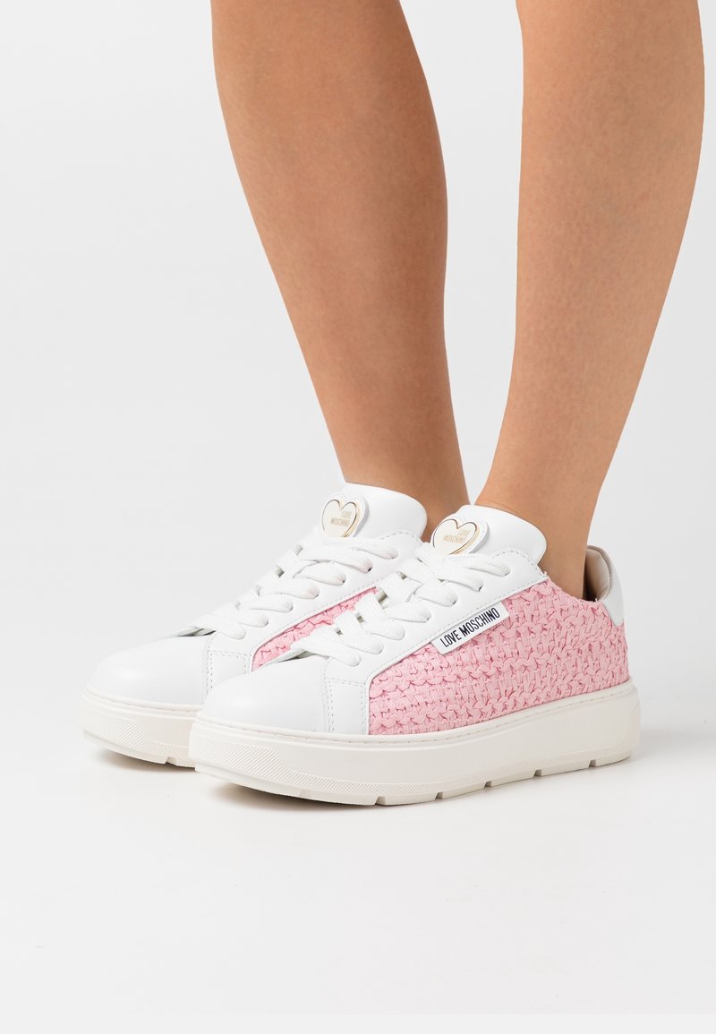 Baskets tissées blanches et roses avec des semelles blanches épaisses portées sur des jambes nues sur un fond blanc uni.