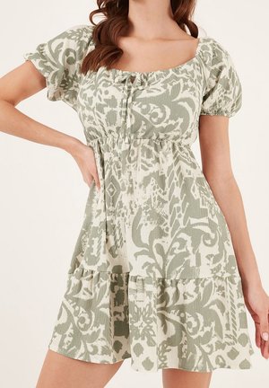 Nainen pukeutuneena lyhythihaiseen, neliökauluksiseen mekkoon, jossa on vihreä ja valkoinen abstrakti kukkakuosi ja laskostettu vyötärö, poseeraa yhdellä kädellä lanteilla.