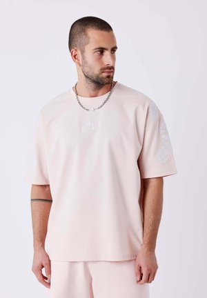Homme aux cheveux courts et barbe portant un t-shirt oversize rose clair et un short assorti, regardant vers sa gauche sur un fond uni.