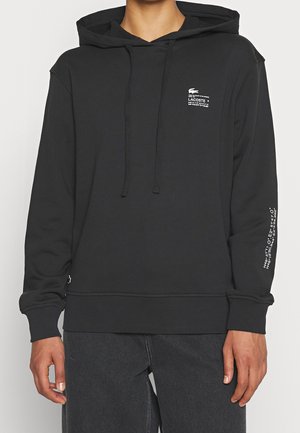 Sudadera negra con bolsillo canguro, capucha con cordón y puños acanalados. Presenta un pequeño logo en el pecho y texto en la manga.