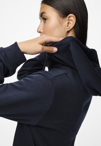 Felpa navy con colletto alto, polsini a costine e texture morbida. Il cappuccio è leggermente tirato indietro, mettendo in mostra i dettagli di design del capo.