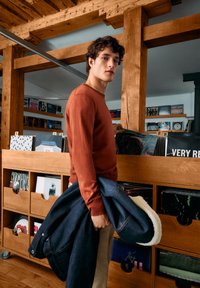 Brown sweater and blue denim jacket with white lining, held by a young man in a wooden interior with vinyl records in the background.
