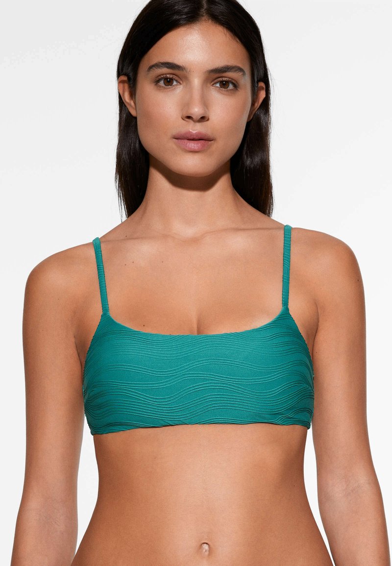 Giovane donna che indossa un top bikini verde con texture e spalline sottili, di fronte a uno sfondo bianco uniforme.