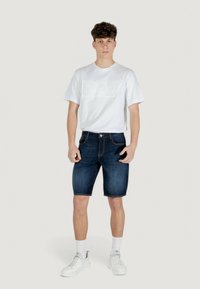 T-shirt blanche à manches courtes avec le logo "EA7" en relief, associée à un short en jean foncé et des baskets blanches. Le modèle se tient debout contre un fond neutre.