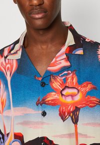 Cămașă cu mânecă scurtă, având un fundal albastru cu modele florale vibrante în nuanțe de roșu și portocaliu, nasturi negri și o textură a materialului moale.
