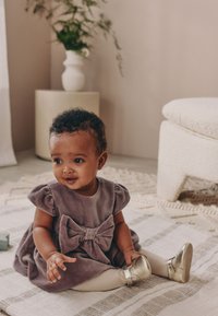 Robe en velours violet avec manches bouffantes et un nœud à l'avant, associée à des collants crème et des chaussures dorées brillantes. Posée sur un tapis à motifs.