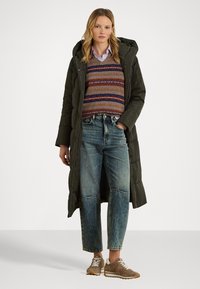 Garšs olīvu zaļš pūkains mētelis virs daudzkrāsaina adīta džempera, apvienots ar augsta vidukļa džinsa biksēm un gaiši brūnām sporta kedām.