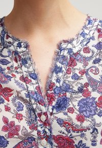 Blommig blus med ett blått och rött mönster på en ljus bakgrund, med en fransad halsringning och knäppning framtill.