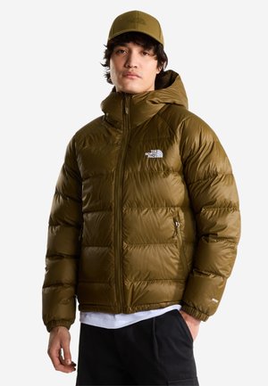 Mann trägt olivgrüne Steppjacke mit Kapuze, weißes T-Shirt, schwarze Hose und passende olivgrüne Kappe vor weißem Hintergrund.