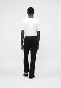 Bijela majica kratkih rukava, crne hlače s elastičnim pojasom i crne tenisice s gumenim đonom, viđene sa stražnje strane na svijetloj pozadini.