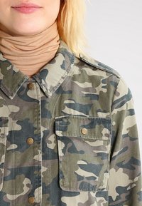 Woman wearing a green and gray camouflage jacket with buttoned flap pockets over a beige turtleneck top.