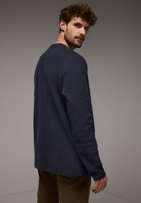 Námořnická košile s dlouhým rukávem z texturované tkaniny, s kulatým výstřihem a ribovanými manžetami, nošená s hnědými kordovým kalhotami.