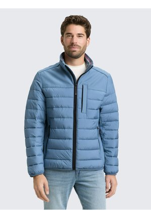 Jeune homme portant une veste matelassée bleu clair avec poches zippées et col montant, associée à un jean bleu clair, sur un fond uni.