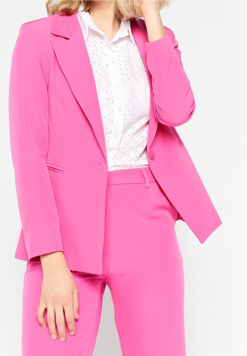 Femme portant un costume rose vif avec un blazer ajusté et un pantalon assorti sur une chemise blanche à motifs œillets boutonnée.