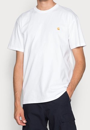 Hombre que lleva una camiseta blanca de manga corta lisa con un pequeño logo amarillo en el pecho izquierdo y pantalones cargo oscuros.