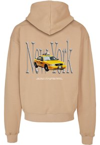 Bezs színű kapucnis felső nagy "New York" felirattal és egy sárga New York-i taxi grafikával a háttérben, alatta kisebb megbízási szöveggel.