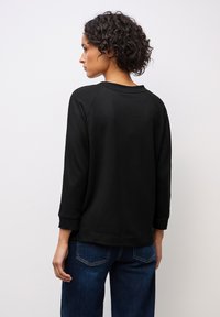 Svart sweatshirt med en avslappnad passform, raglanärmar och rundad halsringning. Mjuk textur, kombinerad med blå denimjeans, sedd bakifrån.