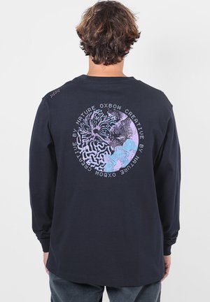 Personne aux cheveux courts et bruns porte un t-shirt noir à manches longues avec un motif abstrait circulaire et un texte dans le dos.