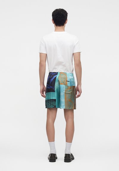 Homme debout, de dos, portant un t-shirt blanc, un short patchwork coloré, des chaussettes blanches et des chaussures noires sur fond uni.
