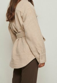 Beige uldne skjortejakke med en blød tekstur, belted talje, knappede manchetter og en afslappet pasform. Båret over mørke bukser.