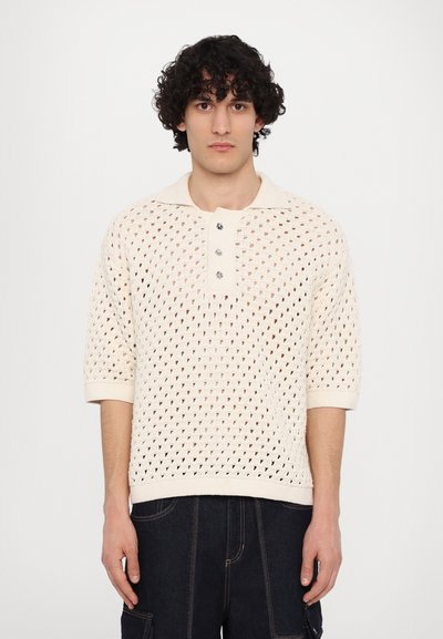 Polo en tricot couleur crème, présentant un motif perforé en diamants, des manches courtes et une patte de boutonnage à trois boutons avec des boutons en argent.