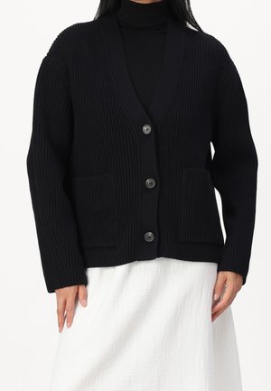 Kvinde iført en sort ribbet cardigan med tre knapper over en sort rullekrave og et hvidt tekstureret skørt.