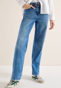 Personne portant un jean droit bleu clair, une chemise blanche, une ceinture noire et des baskets blanches avec des accents verts, debout sur un sol clair.