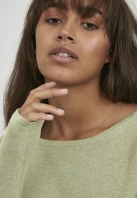 Pull en maille vert clair avec des manches longues et un décolleté arrondi. Texture douce et détails minimaux, porté avec un look naturel.