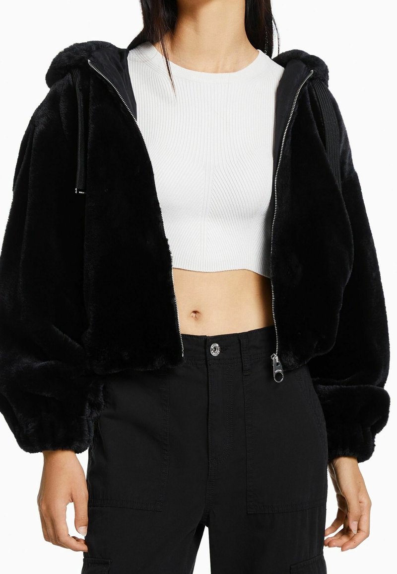Femme portant une veste noire à fermeture éclair en tissu duveteux sur un crop top blanc côtelé et un pantalon taille haute noir, vue du cou jusqu'aux hanches.