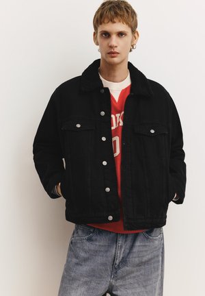 Ung person med kort hår iført en sort knapjakke med en shearling-krave, rød sweatshirt og lyseblå jeans mod en ensfarvet baggrund.