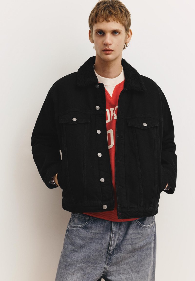 Jeune personne aux cheveux courts portant une veste noire boutonnée avec un col en shearling, un sweat-shirt rouge et un jean bleu clair, sur un fond uni.