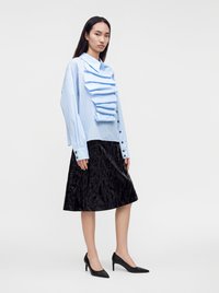 Femme portant un chemisier bleu clair à volants sur le devant, une jupe noire texturée jusqu'aux genoux et des chaussures à talons hauts noires, debout sur un fond blanc.