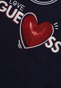 Granatowy sweatshirt z dużą czerwoną cekinową grafiką serca, białym i czerwonym napisem "LOVE GUESS" oraz paskowym wykończeniem kołnierza.